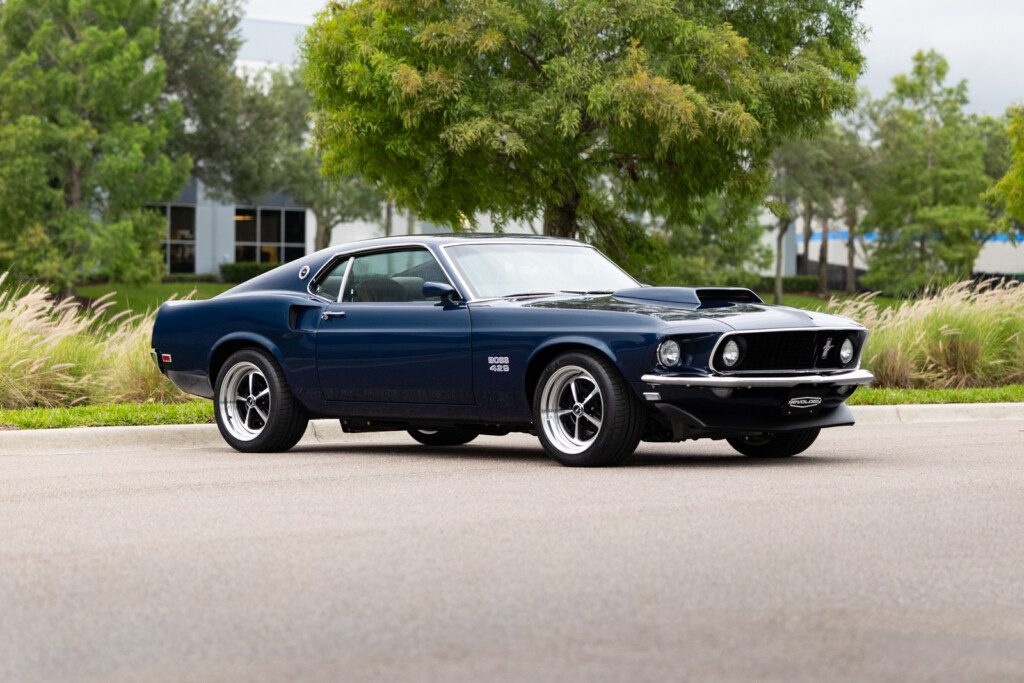 Blue 1969 Revology Mustang Boss 429 Fastback parked on a tree-lined road in a front three-quarter view.